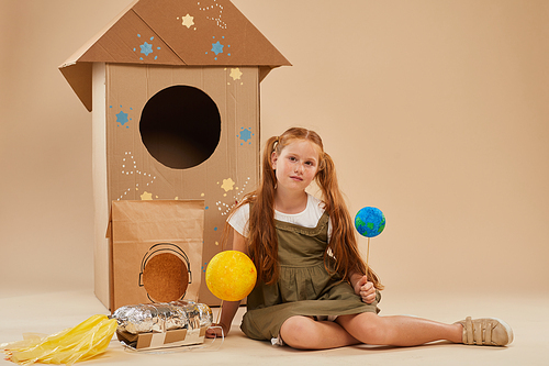 Full length portrait of cute red haired girl playing astronaut while sitting on floor against cardboard rocket in studio, copy space
