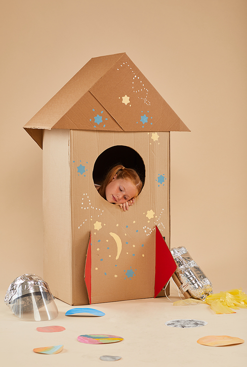 Vertical full length portrait of cute little girl dreaming of space inside cardboard rocket, copy space