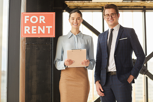 Waist up portrait of two real estate agents smiling at camera while standing next to red FOR RENT sign, copy space