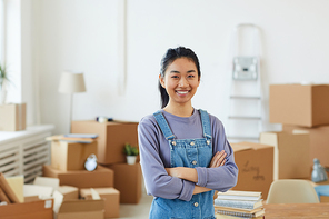 Waist up portrait of cheerful Asian woman smiling at camera while posing in new house with cardboard boxes in background, copy space