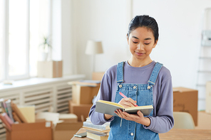 Waist up portrait of young Asian woman writing in planner while standing in new house or apartment with cardboard boxes in background, copy space