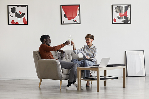 Portrait of young mixed-race couple enjoying champagne while sitting on couch in room decorated by modern art, copy space