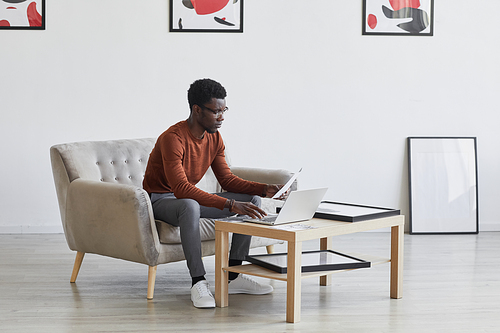 Portrait of young African-American man working at laptop while sitting on couch in room decorated by modern art, copy space