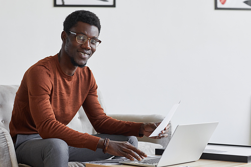 Portrait of contemporary African-American businessman smiling at camera while using laptop and working at home in minimal interior, copy space
