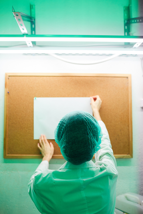 Back view of unrecognizable female doctor pinning blank sign on notice board in laboratory lit by green light