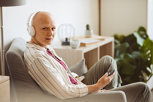 Minimal side view portrait of bald adult woman wearing headphones and looking at camera while listening to music via smartphone in cozy home interior, alopecia and cancer awareness, copy space