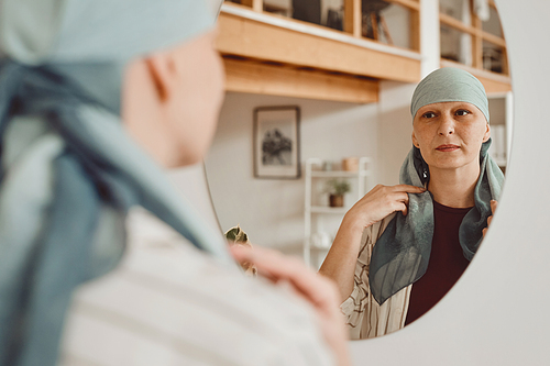 Warm-toned portrait of modern bald woman putting on head scarf while looking in mirror standing in home interior, alopecia and cancer awareness, copy space