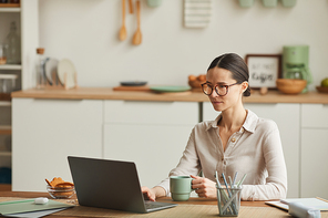 Portrait of elegant young woman drinking coffee while using laptop at cozy home office workplace, copy space