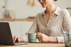 Cropped portrait of smiling young woman drinking coffee while using laptop at cozy home office workplace, copy space