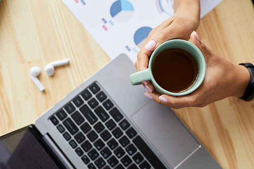 Above view at young woman holding coffee mug over laptop while working at home office, copy space