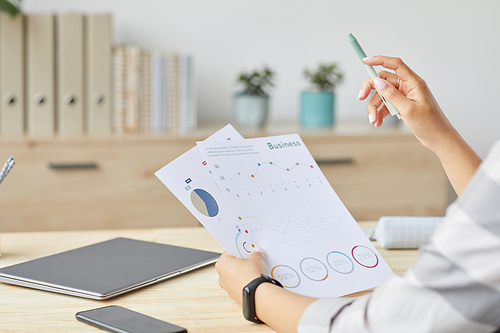 Closeup of unrecognizable young woman holding documents with charts and graphs while sitting by wooden table and working at home office, copy space