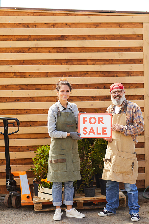 Vertical full length portrait of two contemporary gardeners holding red For Sale sign while standing by plantation outdoors, copy space