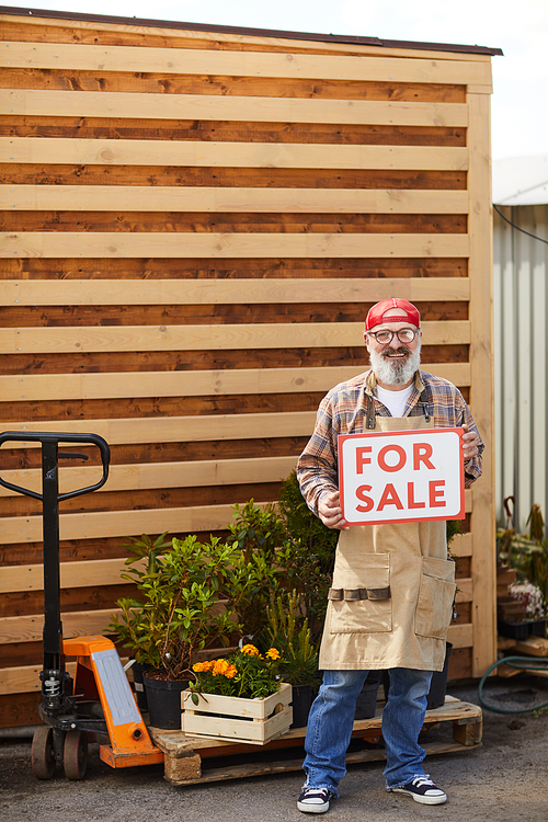 Vertical full length portrait of bearded senior gardener holding red For Sale sign while standing by plantation outdoors, copy space