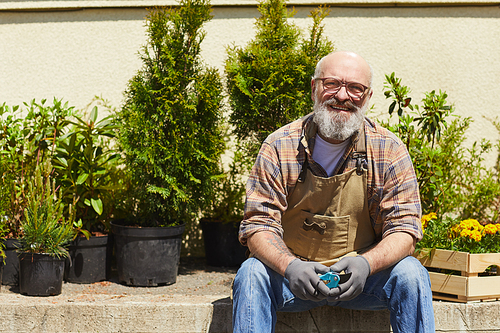 Portrait of smiling senior gardener sitting by potted plants outdoors and looking at camera, copy space