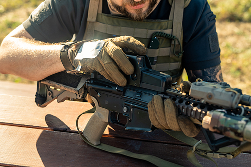 Close-up of bearded man in gloves sitting at table outdoors and assembling rifle