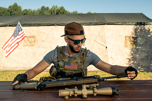 Concentrated young bearded man in sunglasses sitting at wooden table and examining his riffle at American military base