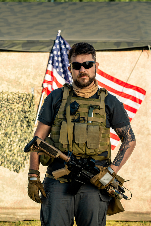 Portrait of serious brutal bearded man in vest with radio device holding riffle against American flag outdoors