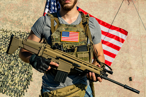 Close-up of bearded American guy in vest with American flag holding personal weapon outdoors