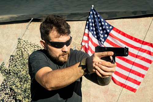 Concentrated American bearded man in sunglasses holding gun and practicing at shooting range