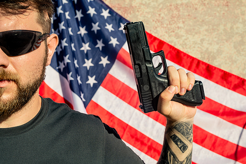 Close-up of brave young bearded man in sunglasses holding gun against American flag