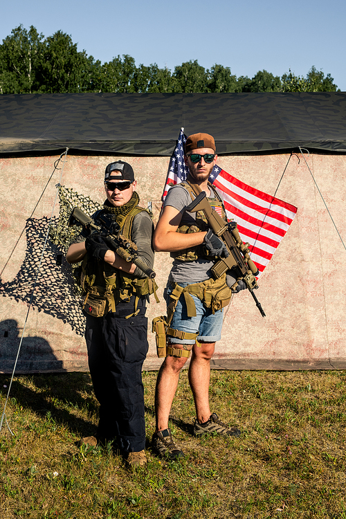 Portrait of serious young friends in caps and sunglasses holding rifles against military tent with American flag