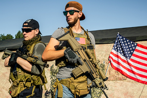 Purposeful young men in sunglasses standing with modern rifles at shooting range, American flag on big tent