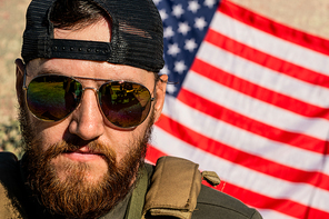 Portrait of serious brutal American man with beard wearing sunglasses and cap standing against national flag
