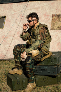 Pensive bearded man in camouflage outfit sitting on metal suitcases and smoking cigarette outdoors