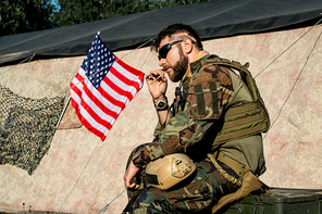 Brutal man in sunglasses smoking cigarette against tent in American military encampment