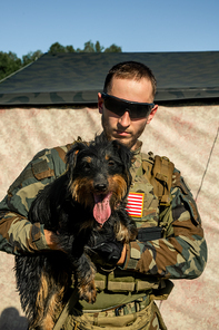Portrait of serious young bearded soldier in sunglasses and camouflage outfit holding cute dog in military encampment