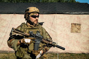 Confident bearded soldier in camouflage outfit and helmet holding rifle and looking into distance