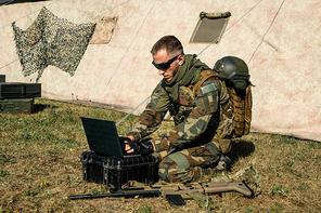 Concentrated young programmer soldier sitting on ground and working with laptop while analyzing army route