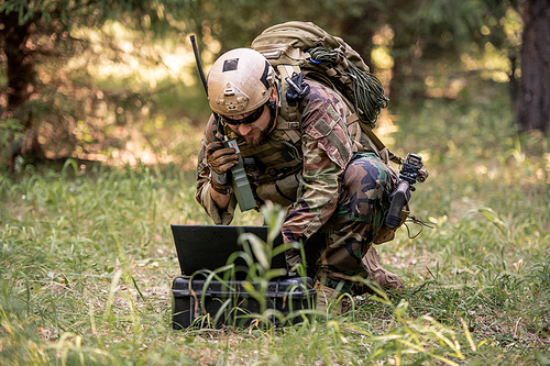 Bearded soldier in camouflage uniform sitting in forest and sending message through radio device while controlling military operation using computer