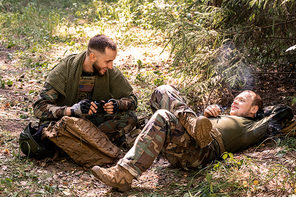 Positive young soldiers in military uniform smoking cigarette and chatting while resting in forest shelter