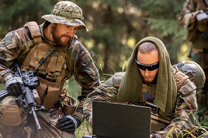 Army American soldiers in camouflage outfits sitting on ground and using laptop while discussing further actions