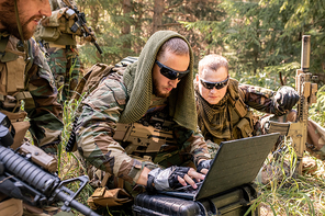 Concentrated army programmer in sunglasses using military computer while getting in touch with military base in forest