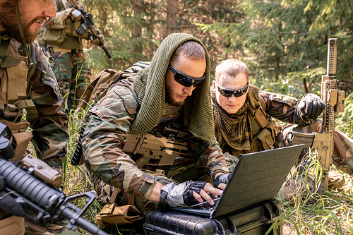 Concentrated army programmer in sunglasses using military computer while getting in touch with military base in forest