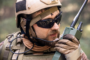 Close-up of bearded soldier in helmet and sunglasses using radio while taking message to colleague during military operation