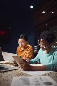 Vertical portrait of two ethnic business people discussing project while working late in dark office, copy space