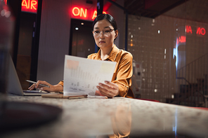 Portrait of Asian businesswoman reading document while working late in dark office, copy space