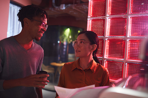 Waist up portrait of two ethnic business people discussing documents while standing outdoors in dim red lighting, copy space