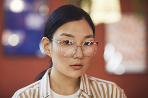 Head and shoulders portrait of young Asian woman wearing glasses looking at camera while posing against red wall in cafe, copy space