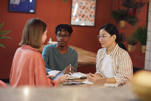 Multi-ethnic group of three young people working on project while sitting at table in cafe against red wall, copy space