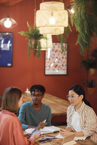Vertical portrait of three young people working on project while sitting at table in cafe against red wall, copy space