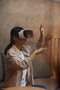 Side view portrait of Asian woman wearing VR gear and gesturing while enjoying immersive experience in futuristic interior