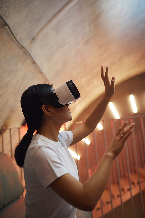 Side view portrait of Asian woman wearing VR gear and gesturing while enjoying immersive reality in futuristic interior