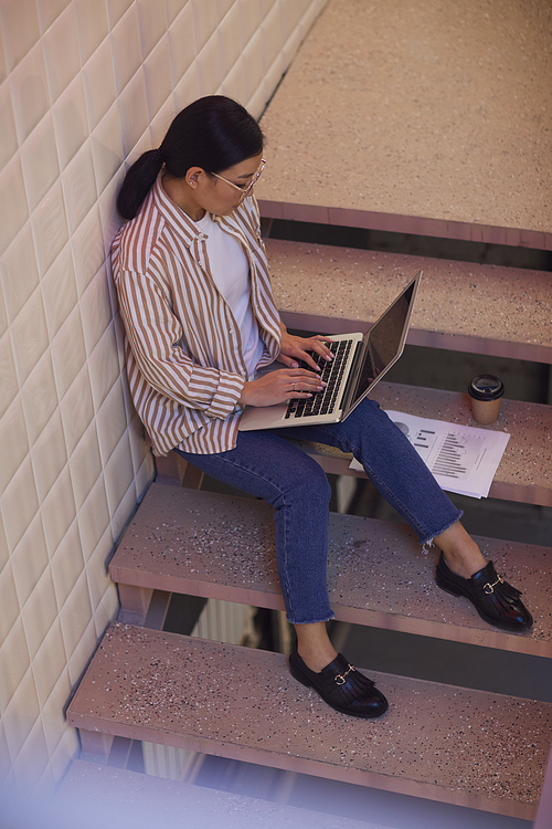 Full length portrait of modern Asian woman using laptop while sitting on staircase by tiled wall