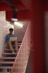 Full length portrait of modern African-American man using laptop while sitting on staircase, copy space