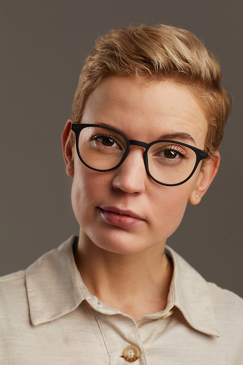 Head and shoulders portrait of modern woman wearing black rimmed glasses with short pixie haircut looking at camera while standing against grey background in studio