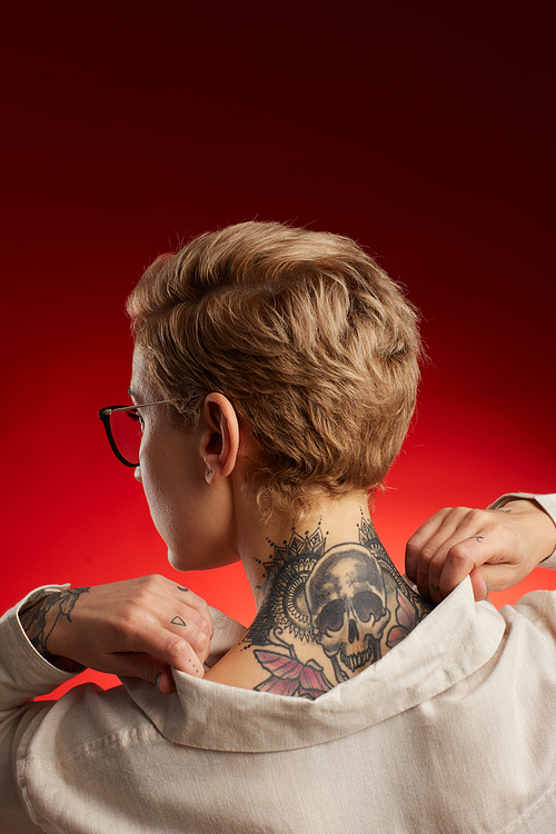 Back view of short haired young woman with neck tattoo featuring scull and butterflies posing against red background in studio